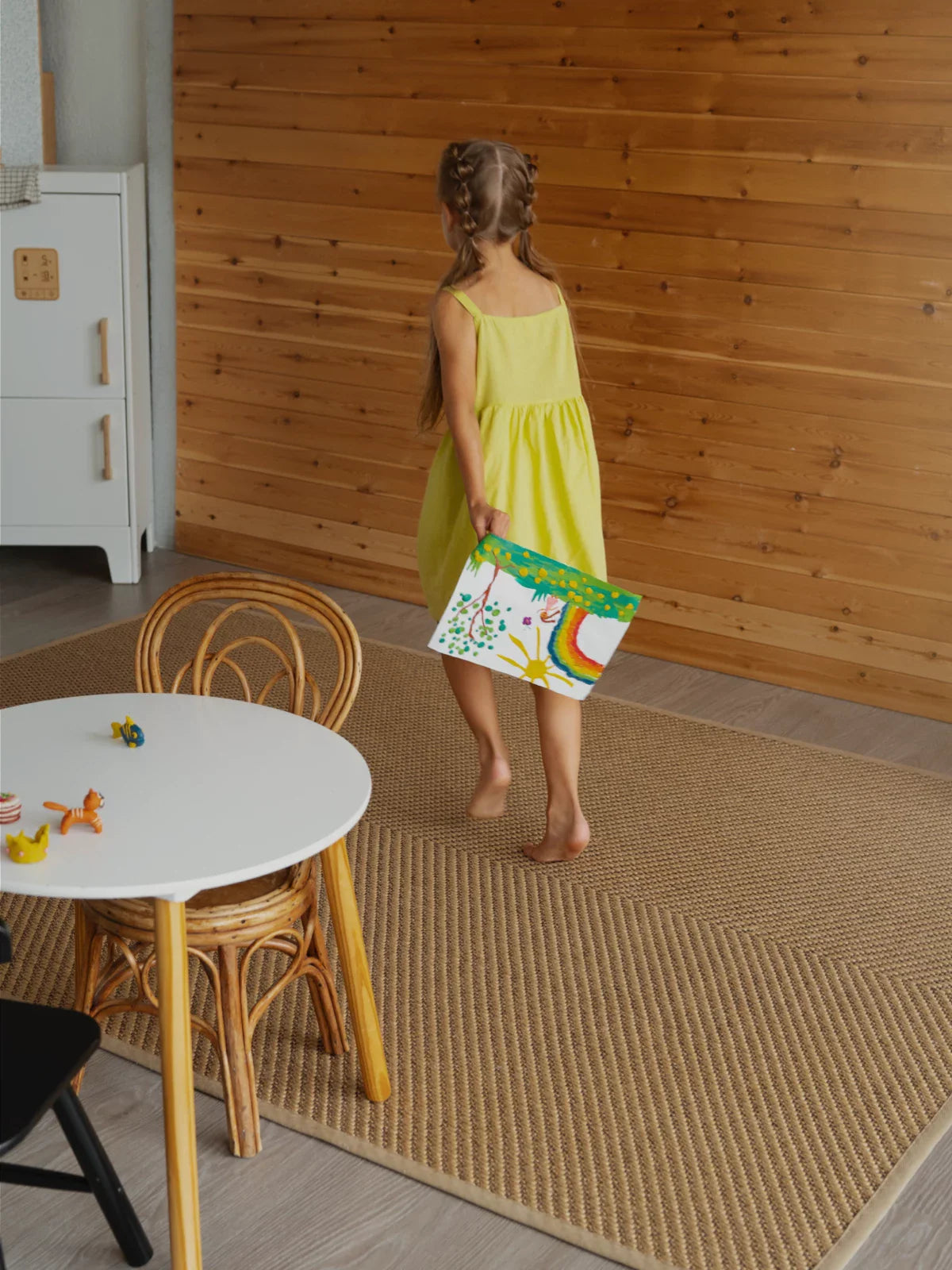 Young girl in a yellow dress walking barefoot on a textured 5x8 outdoor rug in a cozy, wood-accented room, holding a colorful painting.