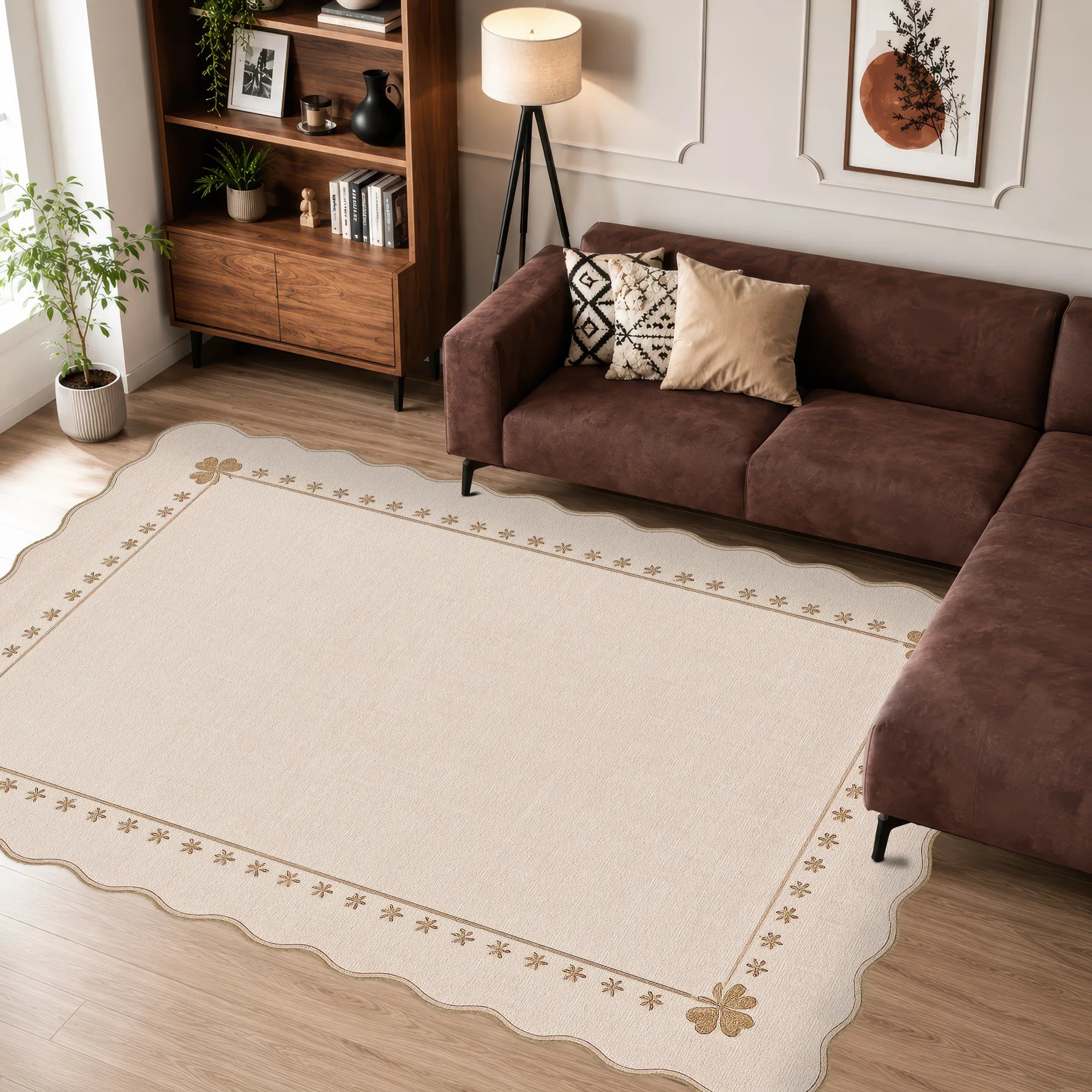 A large cream-colored area rug with a decorative beige border and scalloped edges, shown in a styled living room setting with a brown sectional sofa, wooden bookshelf, and floor lamp.