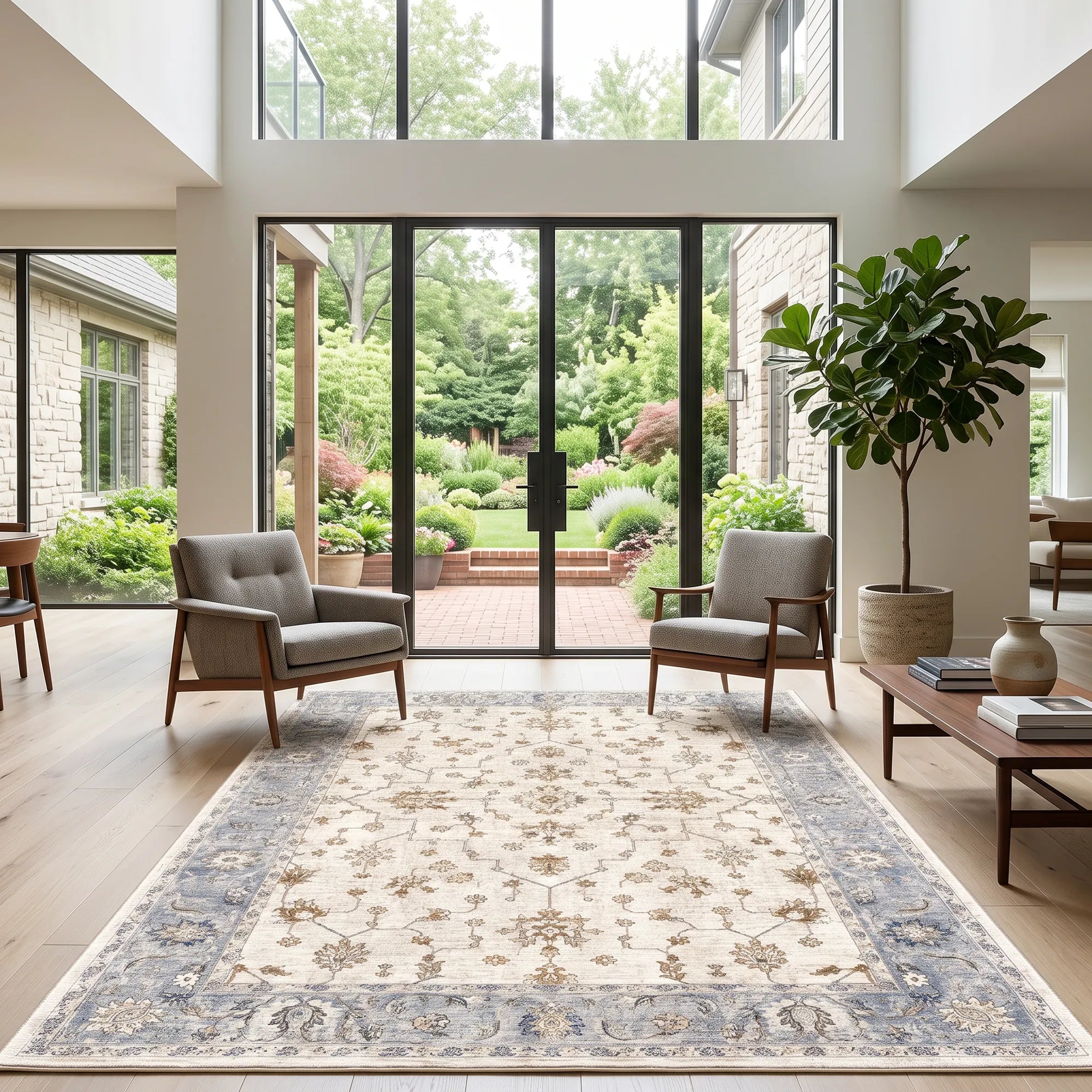 A large rectangular area rug with an ornate traditional border and floral pattern, shown in a bright modern living room with two gray armchairs, a coffee table, and large windows overlooking a garden patio.